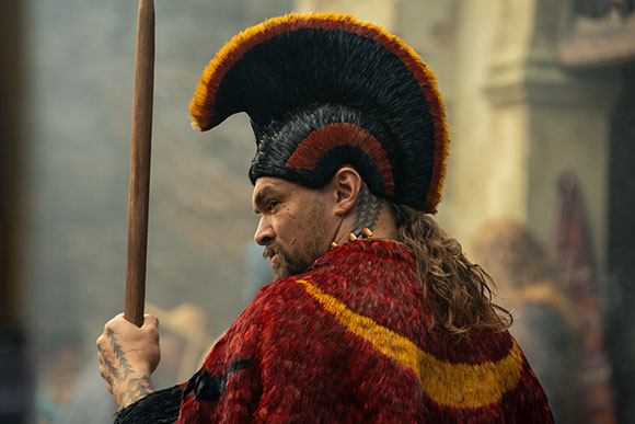 A man with a beard and tattoos wearing a large black and orange feathered headdress and a red patterned cloak.