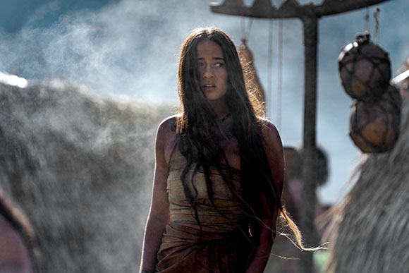 A young woman with long dark hair and tan clothing stands outdoors near thatched structures.
