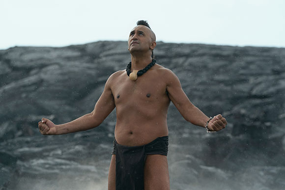 A shirtless man with a dark necklace and dark loincloth stands with arms outstretched against a rocky cliff.