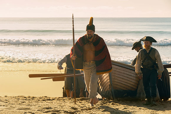 A man in a patterned cloak walks on a sandy beach holding a staff near a wooden boat and other men.