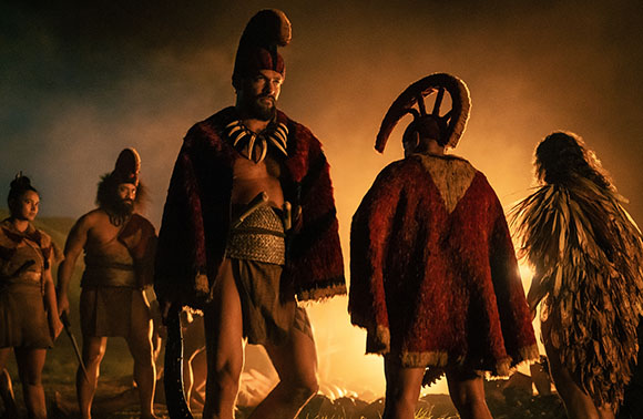 Five people in feathered and fur tribal clothing stand around a bright fire at night.
