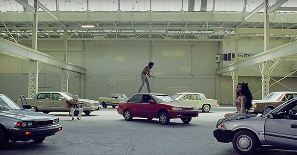 A man stands on a red car in a large warehouse surrounded by several other parked cars and people.