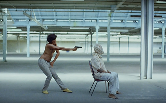 A shirtless man aims a handgun at a person sitting in a chair in a large, empty warehouse.