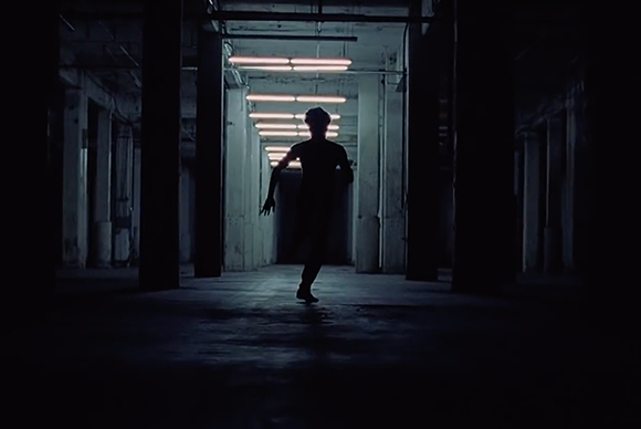 Silhouette of a person dancing in a dark, industrial corridor under rows of fluorescent lights.