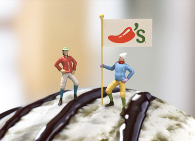 Two tiny figures stand on chocolate-drizzled ice cream next to a flag with a red chili pepper logo.