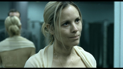 A blonde woman in a light-colored top looks to the side in a dimly lit room.