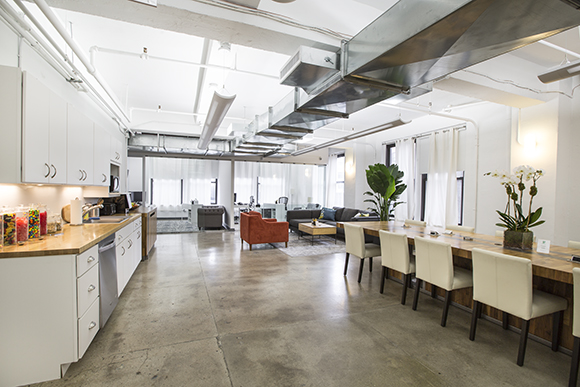 A bright, open-concept loft with a white kitchen, long wooden dining table, and industrial ceiling ducts.