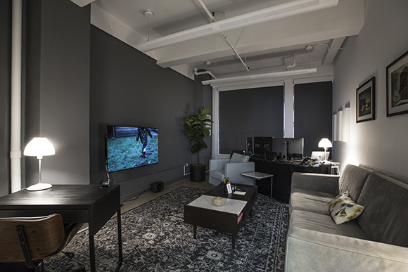 A living room featuring a gray sofa, a dark desk with a lamp, a television on a gray wall, and a patterned rug.