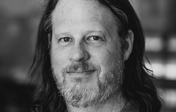 Black and white close-up portrait of a man with long hair and a salt-and-pepper beard looking at the camera.