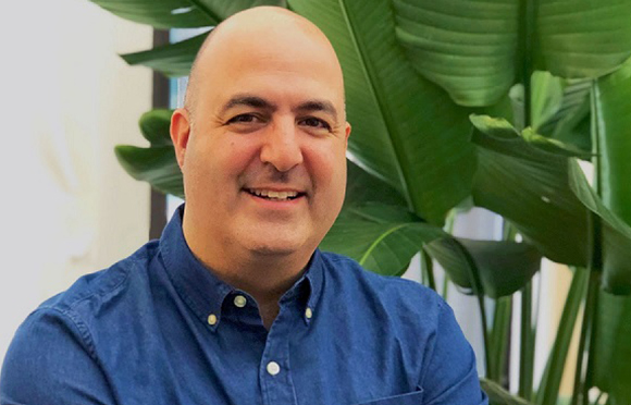 A smiling bald man wearing a blue button-down shirt in front of large green tropical leaves.
