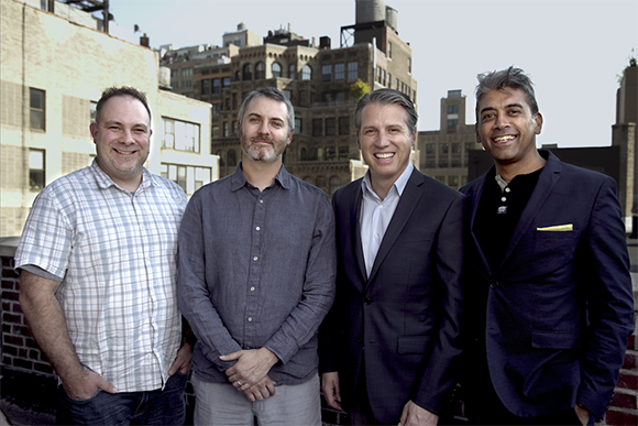 Four men standing in a row smiling, with urban buildings in the background.