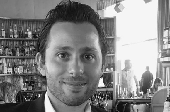 Black and white close-up portrait of a smiling man in a bar with shelves of bottles in the background.