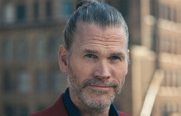Close-up portrait of a middle-aged man with grey hair in a bun and a salt-and-pepper beard looking at the camera.