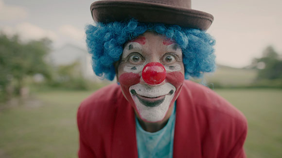 A person wearing clown makeup, a bright blue curly wig, a brown hat, and a red jacket.