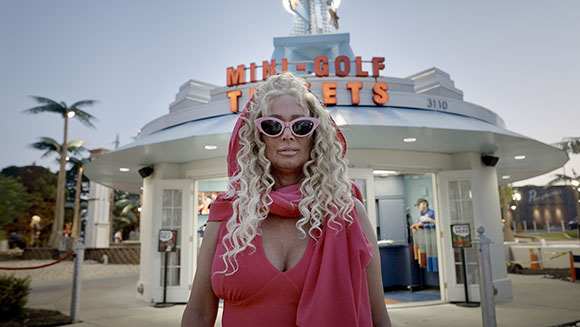 A woman with long blonde curls and pink sunglasses stands before a mini-golf building at dusk.
