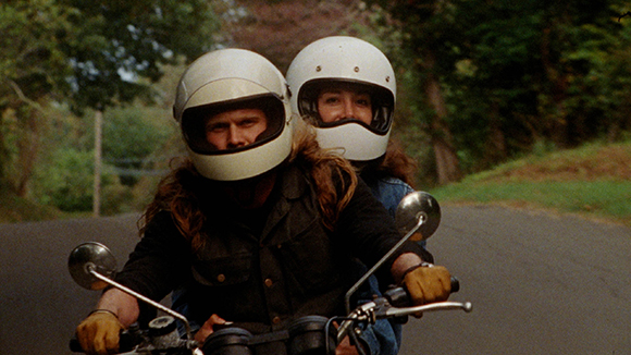 Two people with long hair wearing white helmets ride a motorcycle down a road.