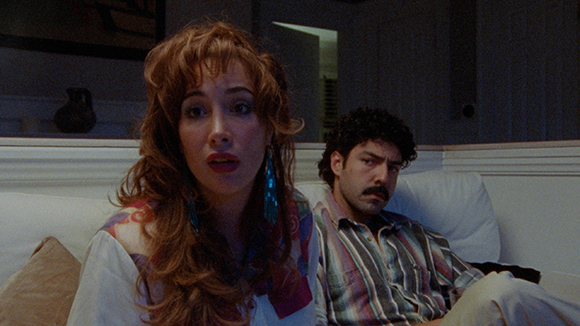A woman with wavy reddish hair looks forward with a surprised expression while a man sits behind her.