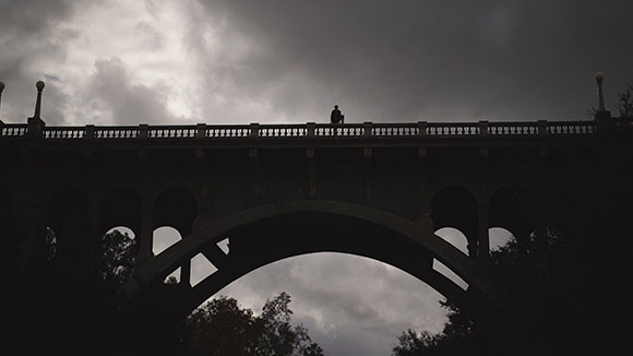 Silhouette of a large arched bridge with a single person standing on the railing against a cloudy sky.