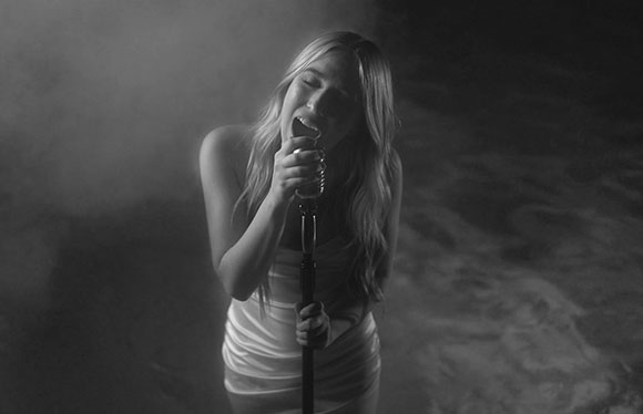 Black and white photo of a woman in a white dress singing into a vintage microphone amidst smoke.