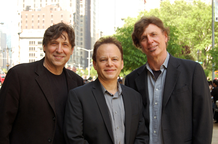 Three men in dark blazers and collared shirts smiling for a portrait outdoors.