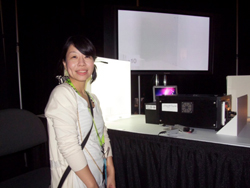 A woman smiles at the camera in front of a table with electronic devices and a large screen displaying a bright white image.
