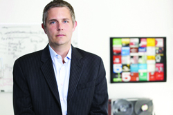 A man wearing a dark pinstripe blazer and light blue button-down shirt stands in an office setting.