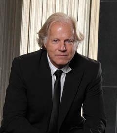 A man with light hair wearing a black suit, white shirt, and black tie looking at the camera.