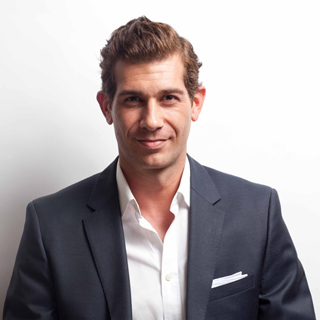 A man with wavy brown hair wearing a dark blazer over an open-collared white shirt.