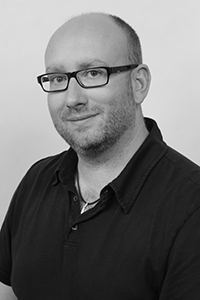 Black and white headshot of a man with glasses and a short beard wearing a dark polo shirt.