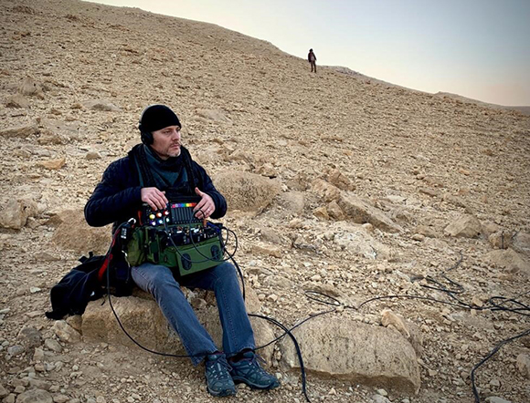 A man in a black beanie and blue jacket sits on a rocky slope using a sound mixing board with various cables.