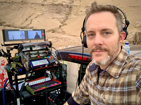 A man with a beard and headphones smiles next to a mobile audio mixing station in a desert landscape.