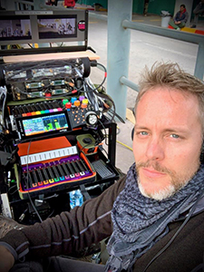 A man with a beard and scarf sits beside a cart filled with monitors, controllers, and electronic gear.