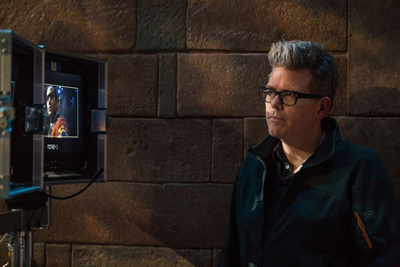 A man with glasses looks toward a film monitor displaying a scene of a person in a red uniform.