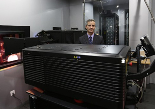 A man in a suit stands behind a large, black, rectangular piece of industrial equipment in a laboratory.