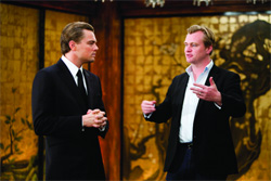 A man in a black suit and tie listens to a man in a black blazer and light blue shirt gesturing with his hands.