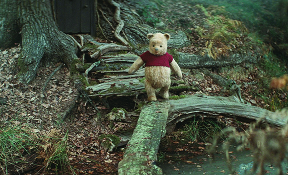 A small brown teddy bear wearing a red shirt sits on a mossy log in a wooded area.