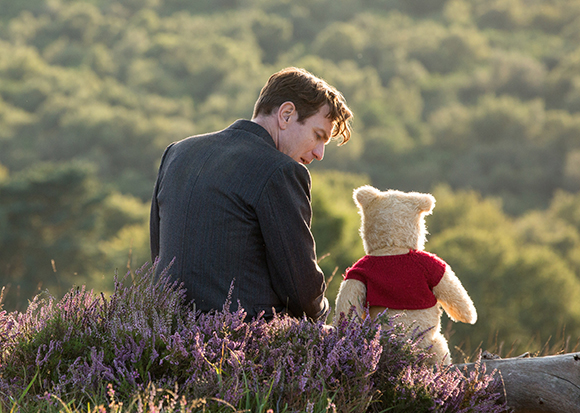 A man in a dark suit sits next to a teddy bear wearing a red sweater in a field of purple heather.