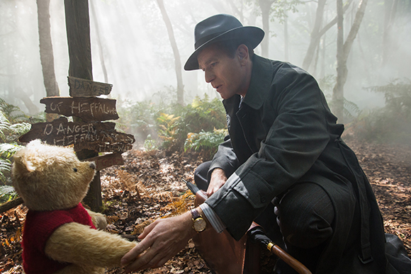 A man in a dark coat and hat crouches in a misty forest next to a small teddy bear.