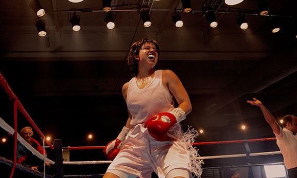 A female boxer in a white tank top and shorts cheers with her mouth open in a boxing ring.