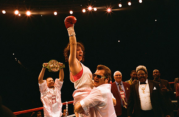 A female boxer raises a red glove in victory while being lifted by a man in a boxing ring.