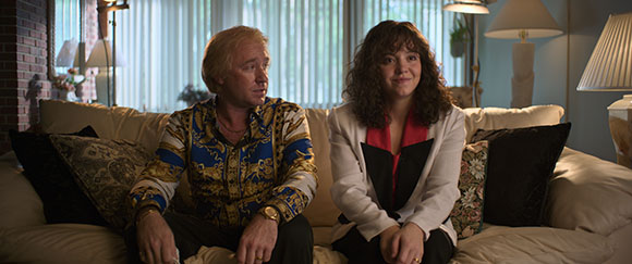 A man in a patterned blue shirt and a woman in a white blazer sit together on a beige sofa.