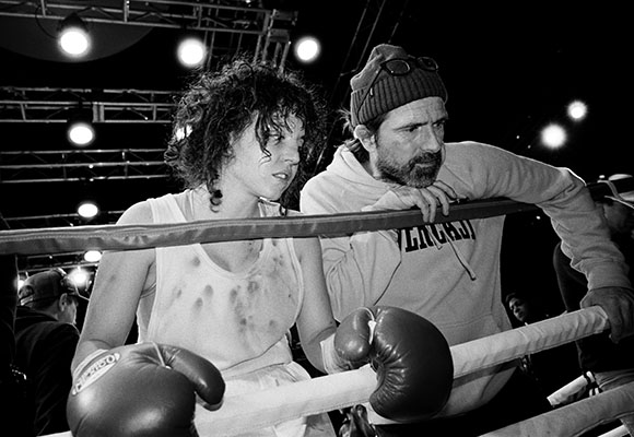A female boxer in a tank top and her bearded trainer lean over the ropes of a boxing ring.