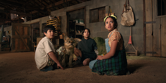 Three children and a small, furry creature sit on the dirt floor of a dimly lit barn.