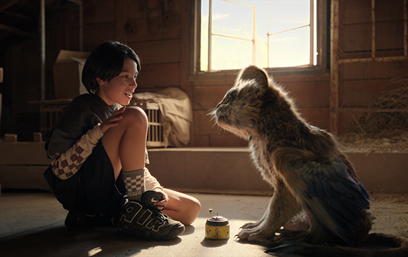 A young person sits on a wooden floor, smiling at a small, winged feline creature.