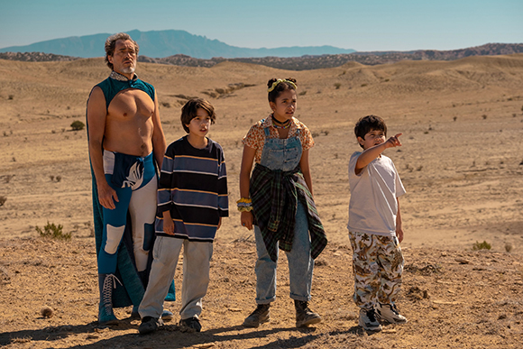A man in a blue cape and three children stand in a vast, sandy desert under a clear sky.
