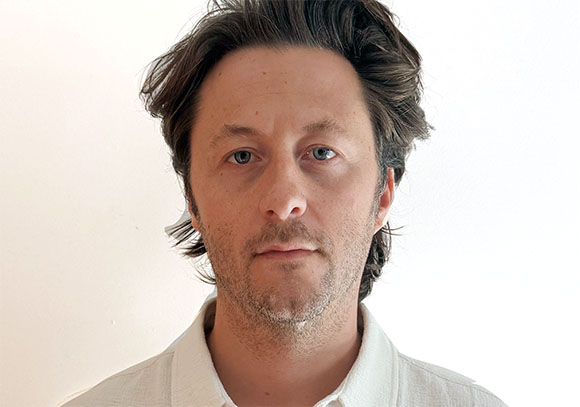 A man with wavy dark hair and blue eyes looking directly at the camera against a white background.