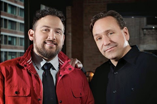 Two men smiling, one in a red jacket and tie, the other in a black button-down shirt.