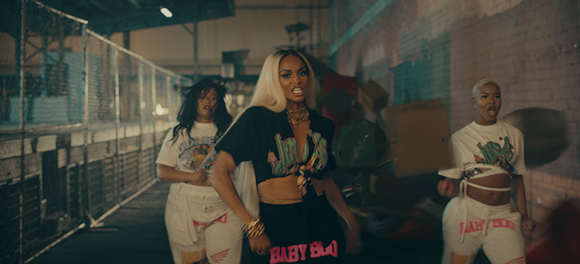 Three women in stylish streetwear posing in a warehouse with industrial shelving and brick walls.