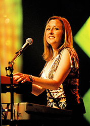 A smiling woman with blonde hair plays a keyboard and sings into a microphone on a stage.