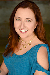 A smiling woman with long reddish-brown hair wearing a blue top and gold necklaces.